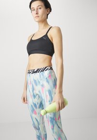 Fekete sportmelltartó vékony pántokkal, világoskék és rózsaszín mintás leggings-szel párosítva. Zöld vízpalackot tart a kezében. Minimalista dizájn.