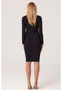 Czarna dopasowana sukienka z długim rękawem, sięgająca do kolan, wykonana z gładkiego materiału, z detalami szwów; noszona z cielistymi szpilkami.