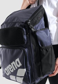 Mørkeblå rygsæk med flere lynlåslommer, mesh-detaljer og et stort hvidt "arena"-logo på siden. Struktureret stof.
