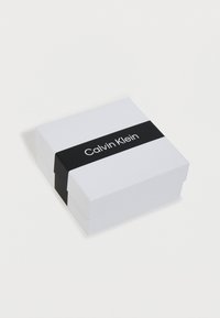 Caja de regalo de Calvin Klein, blanca con una banda negra que presenta el logotipo. Forma rectangular, superficie lisa, diseño minimalista.
