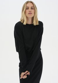 Fekete, túlméretezett, hosszú ujjú felső, bordás mandzsettával, fitted fekete szoknyával párosítva. Puha textúra, minimalista design, látható minták nélkül.