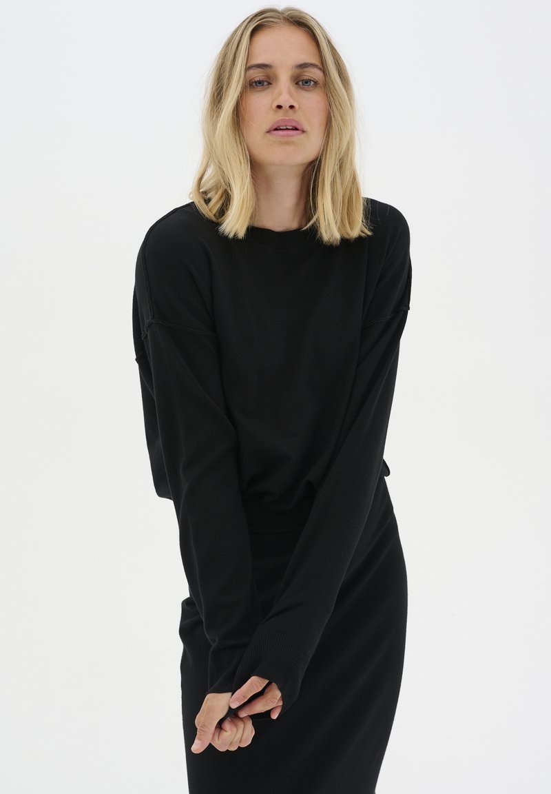 Fekete, túlméretezett, hosszú ujjú felső, bordás mandzsettával, fitted fekete szoknyával párosítva. Puha textúra, minimalista design, látható minták nélkül.