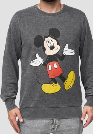 Mężczyzna noszący szarą bluzę z grafiką Mickey Mouse w stylu vintage, prezentującą gest wzruszania ramionami, w czerwonych szortach i żółtych butach.