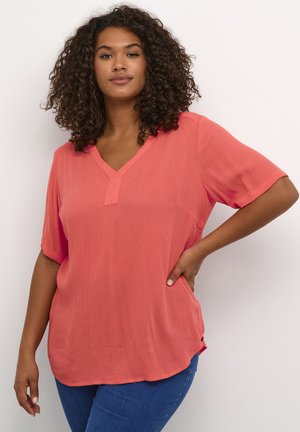 Blouse corail à col en V avec manches courtes, en tissu texturé et fentes sur les côtés, associée à un jean bleu. Surface lisse et coupe décontractée.