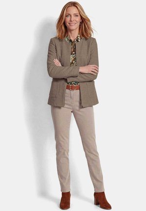 Vrouw met schouderlang blond haar staat met gekruiste armen, draagt een bruine jas, een patroon overhemd, beige broek en bruine enkellaarsjes.