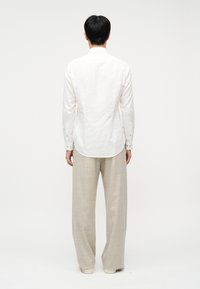 Άνδρας με λευκό μακρυμάνικο πουκάμισο και μπεζ φαρδιά παντελόνια, γυρισμένος πλάτη, μπροστά σε μια απλή λευκή φόντο.