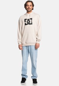 Beige hoodie with large black DC logo on front, featuring a kangaroo pocket and adjustable drawstring hood. Paired with light blue jeans and black sneakers.