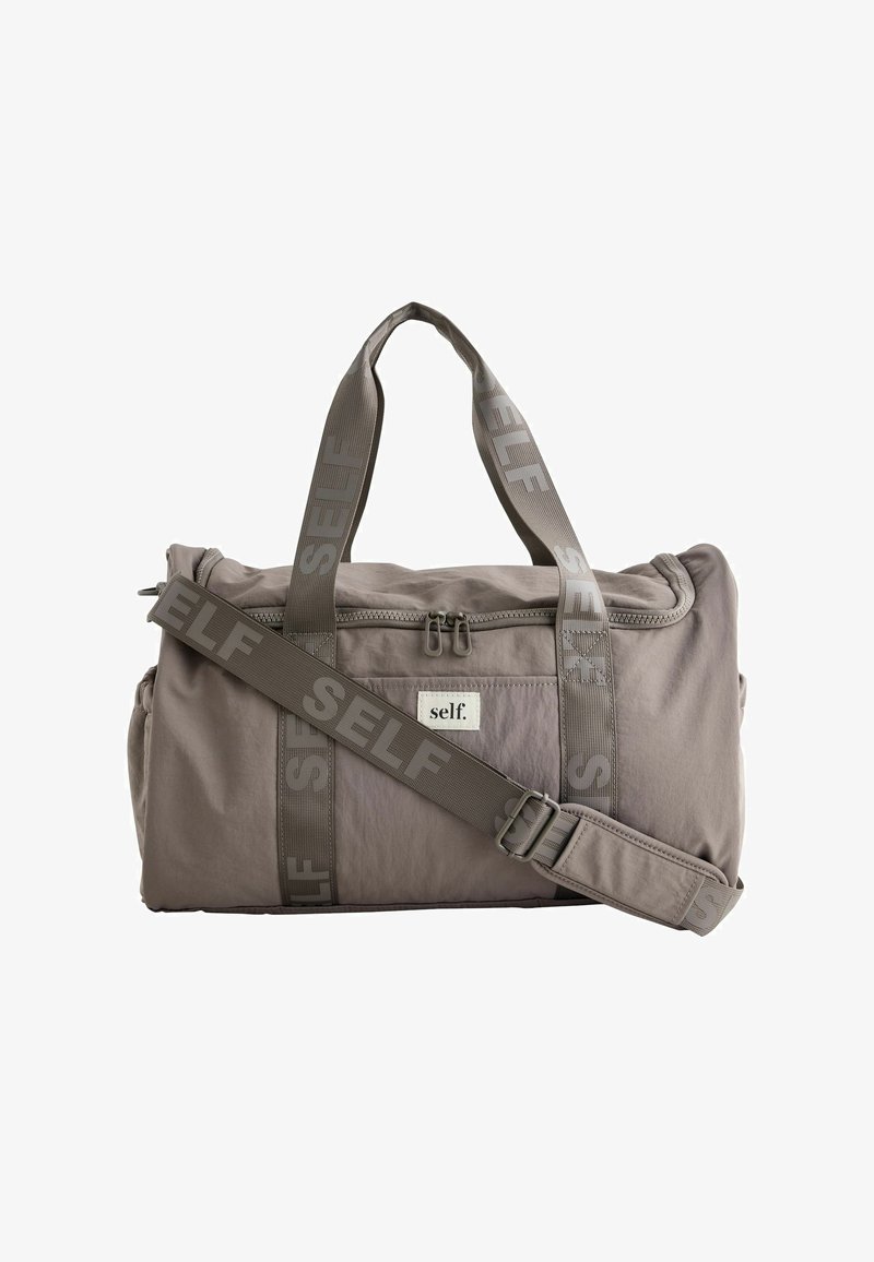 Lysegrå duffelpose med to håndtag og en justerbar skulderrem, begge med "SELF"-logo trykt, og en lynlåslomme foran.