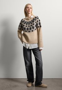 Beige Strickpullover mit schwarzem und blauem Blumenmuster, Rippabschluss. Über einem weißen Hemd mit Kragen getragen und kombiniert mit dunklen Jeans und beigen Schuhen.