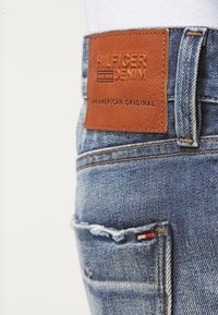 Siniset farkut, joissa on ruskea nahkainen merkki, johon on painettu "HILFIGER DENIM". Taskuissa kulutuksen jälkiä ja pieni punavalkoinen ja sininen koriste.