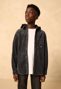 Gray corduroy shirt with a hood, featuring a front pocket and button closure, worn over a white T-shirt, paired with dark jeans.