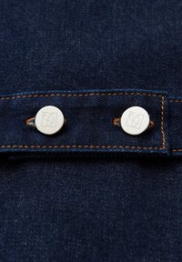 Tessuto in denim scuro con due bottoni bianchi incisi con loghi, cuciture arancioni a contrasto che evidenziano i dettagli delle cuciture.