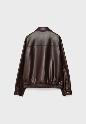 Femme portant une veste en cuir marron, un top court blanc, un jean à jambes larges et un long collier de perles, debout sur un sol en béton.