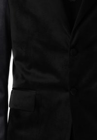 Svart kavaj i slätt tyg, med en enda knappstängning, notch-revers och bröstficka. Texturerad ytdetalj är synlig.