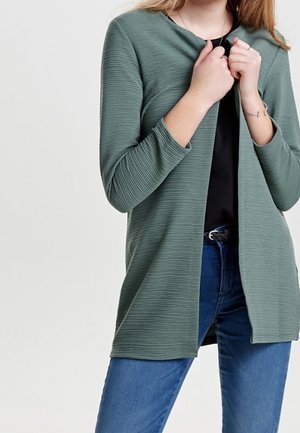 Frau trägt eine grüne, gerippte, offene Strickjacke über einem schwarzen Top und blauen Jeans und richtet die Strickjacke mit beiden Händen.