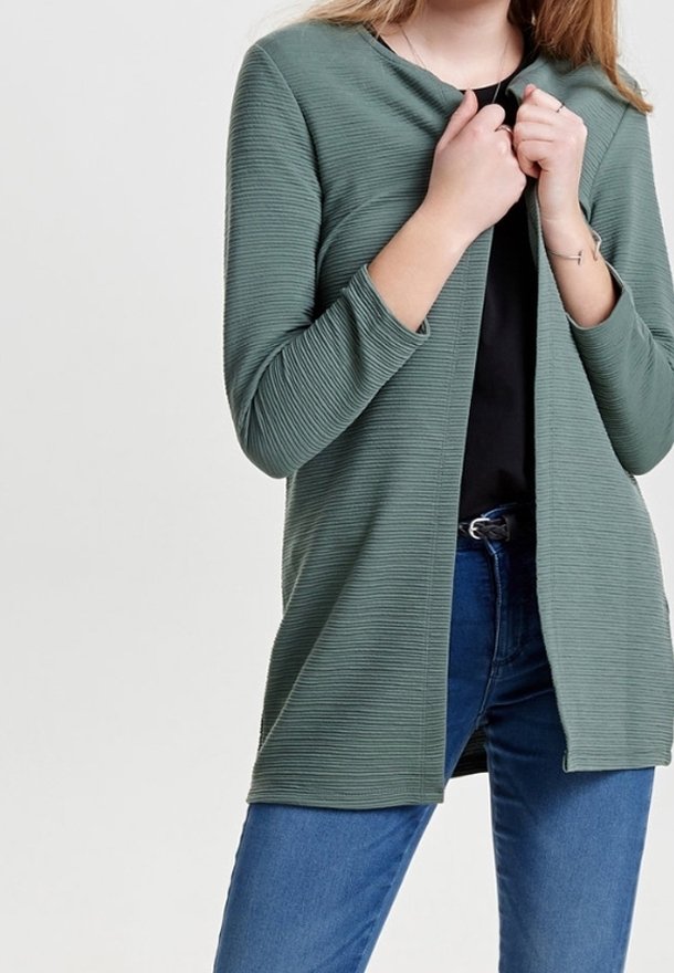 Femme portant un cardigan vert côtelé à ouverture devant, sur un haut noir et un jean bleu, ajustant le cardigan avec ses deux mains.
