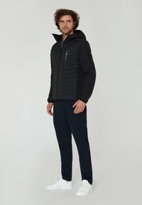 Sort pufferjakke med lynlås foran, hætte og sidelommer. Har stitched chevron mønstre og kontrasterende sorte ærmer.