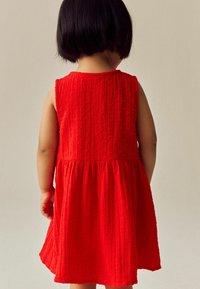 Röd ärmlös klänning med en texturerad tyg som har små upphöjda prickar, rynkad vid midjan och som växer ut mot fållen.