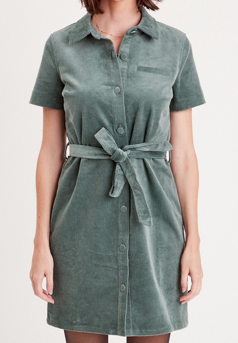 Abito camicia in corduroy verde con maniche corte, apertura con bottoni frontale e cintura annodata in vita. Presenta una tasca sul petto e costine verticali.
