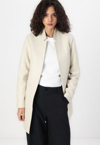 Cappotto in misto lana beige con colletto alto, apertura frontale e tasche laterali. Indossato sopra una camicia bianca e pantaloni neri a vita alta.