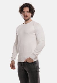 Maglione bianco a maniche lunghe, con texture, colletto a giro e polsini a costine, abbinato a jeans grigio scuro, su uno sfondo neutro.