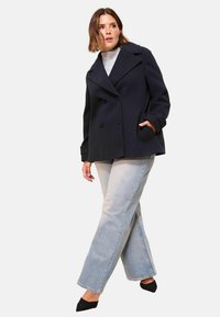 Femme portant un manteau noir à double boutonnage, un pull col roulé gris, un jean bleu clair et des chaussures noires à talons pointus, se tenant avec les jambes croisées.