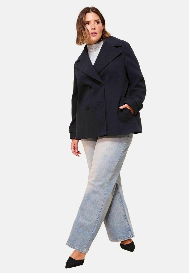 Femme portant un manteau noir à double boutonnage, un pull col roulé gris, un jean bleu clair et des chaussures noires à talons pointus, se tenant avec les jambes croisées.