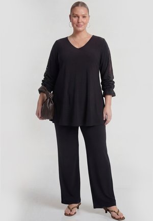 Frau trägt schwarzen V-Ausschnitt Top mit Spitzenärmeln, schwarze weite Hose, Leopardenmuster-Absätze, hält eine dunkelbraune Handtasche.