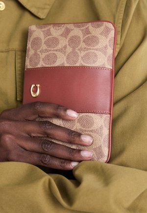 Donkere hand die een beige en bruin patroonportemonnee met gouden logo vasthoudt, tegen een olijfgroene stofachtergrond.