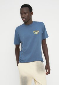 Μπλε βαμβακερό t-shirt με κοντά μανίκια και στρογγυλή λαιμόκοψη, με κίτρινο και μπλε λογότυπο "Rip Curl" στο στήθος.