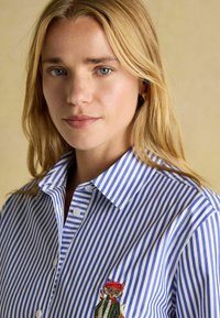 Modrá a biela pruhovaná košeľa s golierom na gombík, s vyšitým karikatúrnym líškom v červenej čiapke a zelenej bunde na ľavej strane hrudníka.