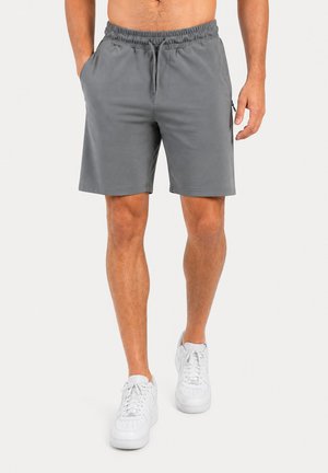 Homme portant un short de sport gris avec cordon de serrage et poche zippée latérale, associé à des baskets blanches, debout devant un fond uni.