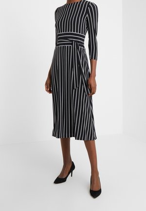 Woman wearing a black midi dress with white vertical stripes, three-quarter sleeves, a tied waistband, and black high-heeled shoes.