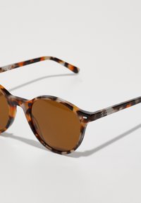 Tortoiseshell päikeseprillid ümmarguste läätsede, pruuni tooniga läätsede ja õhukeste raskustega. Küljel on brändi logo. Sile tekstuur.