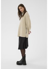 Pull en beige à côtes avec fentes sur les côtés, associé à une jupe satin noire et des bottines en cuir noires chunky, dotées d'une semelle texturée.