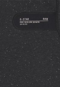 Black textured fabric with white speckles, featuring a rectangular black label with "G-STAR RAW" and production details.