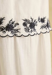 Biely textil s prepracovanou čiernou kvetinovou výšivkou pozdĺž lemu, zakončený ozdobným vlnkovaným okrajom, ľahký materiál s textúrou.