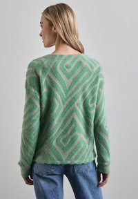 Kobieta o blond włosach, ubrana w zielono-beżowy sweter z geometrycznym wzorem i niebieskie dżinsy, widziana od tyłu na jednolitym tle.