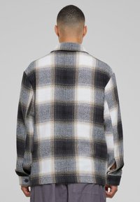 Mężczyzna w czarnej, białej i beżowej kratkowanej marynarce, pokazany z tyłu na jednolitym szarym tle.