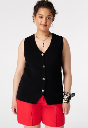 Vrouw met krullend haar draagt een zwarte mouwloze top met knopen, rode shorts, een gouden ketting, creool oorbellen en een armbandsjaal met luipaardprint.