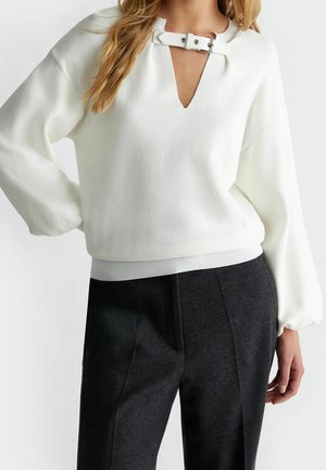 Kvinde iført en hvid sweater med spænde ved V-halsen og sorte skræddersyede bukser, poserer med den ene hånd på hoften.