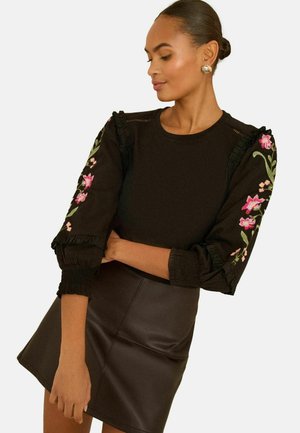 Fekete pulóver fodros, hímzett virágmintás ujjal; fekete műbőr mini szoknyával párosítva; sima textúra, testhezálló sziluett.