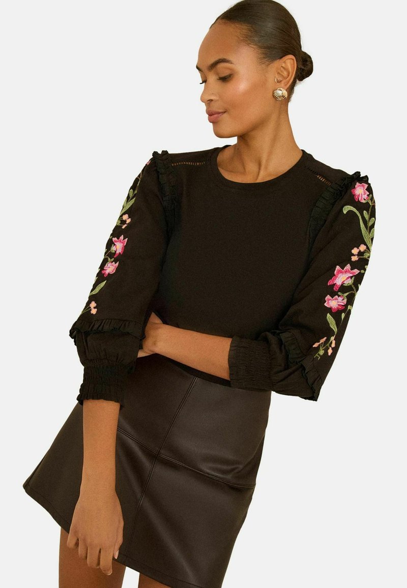 Fekete pulóver fodros, hímzett virágmintás ujjal; fekete műbőr mini szoknyával párosítva; sima textúra, testhezálló sziluett.