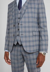 Completo grigio a quadri con design slim fit e dettagli in blu. Comprende un blazer e pantaloni con pettorina. Dettagli dei bottoni visibili.