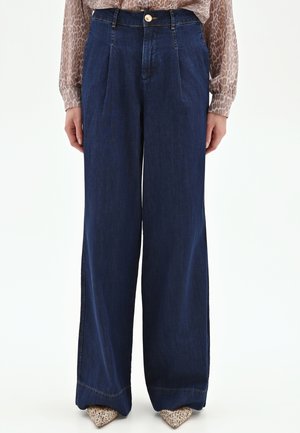 Donna che indossa pantaloni di denim blu scuro a vita alta e gamba larga, abbinati a una camicetta con fantasia e scarpe appuntite con texture.