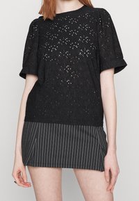Mujer con blusa de encaje negra de manga corta y falda corta de rayas negras, de pie contra un fondo claro liso.
