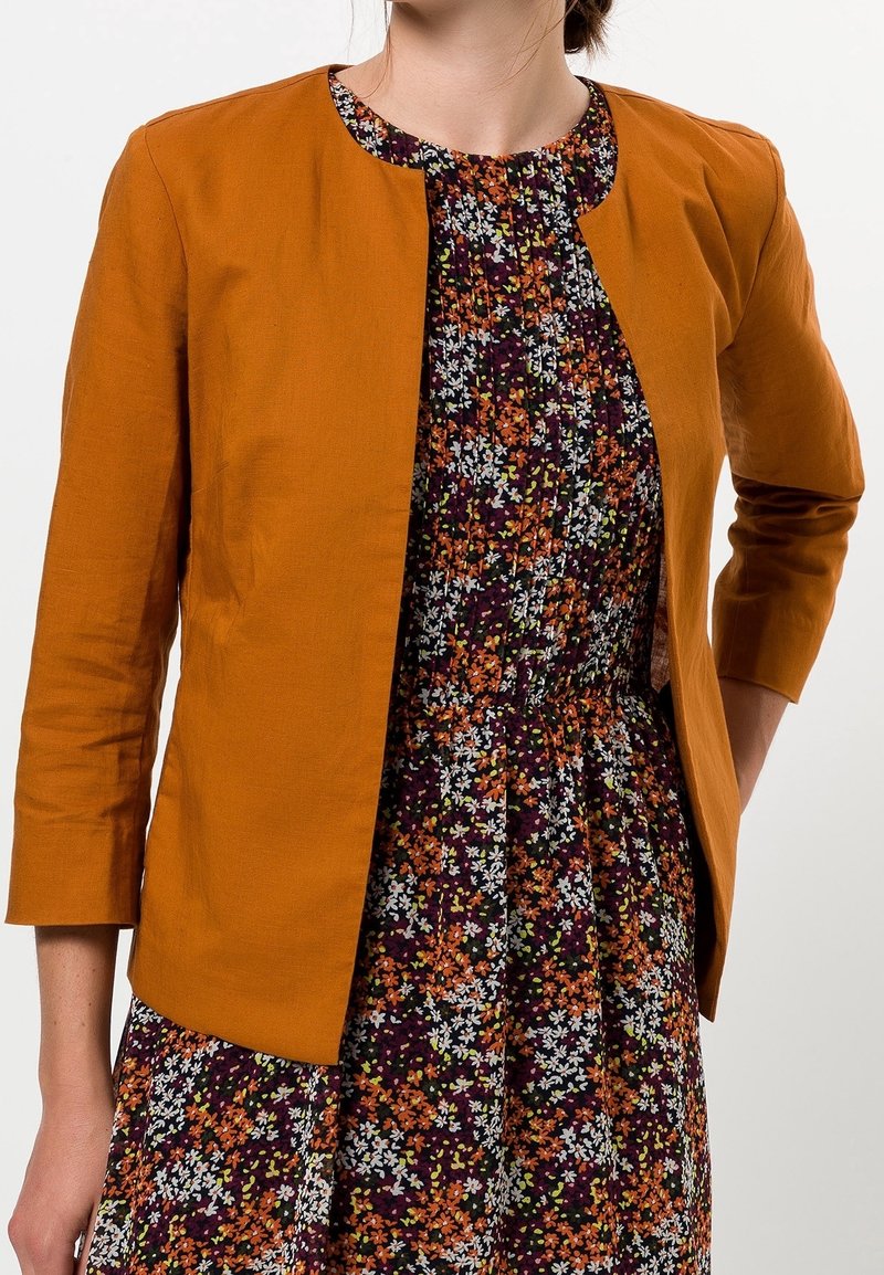 Mujer viste una chaqueta abierta de color mostaza sobre un vestido floral oscuro con pequeñas flores de color naranja, blanco y verde.