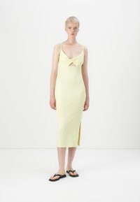 Robe midi en tricot de couleur jaune clair, avec un design en torsion au niveau de la poitrine, des bretelles fines et une fente sur le côté. Le modèle porte des tongs noires.