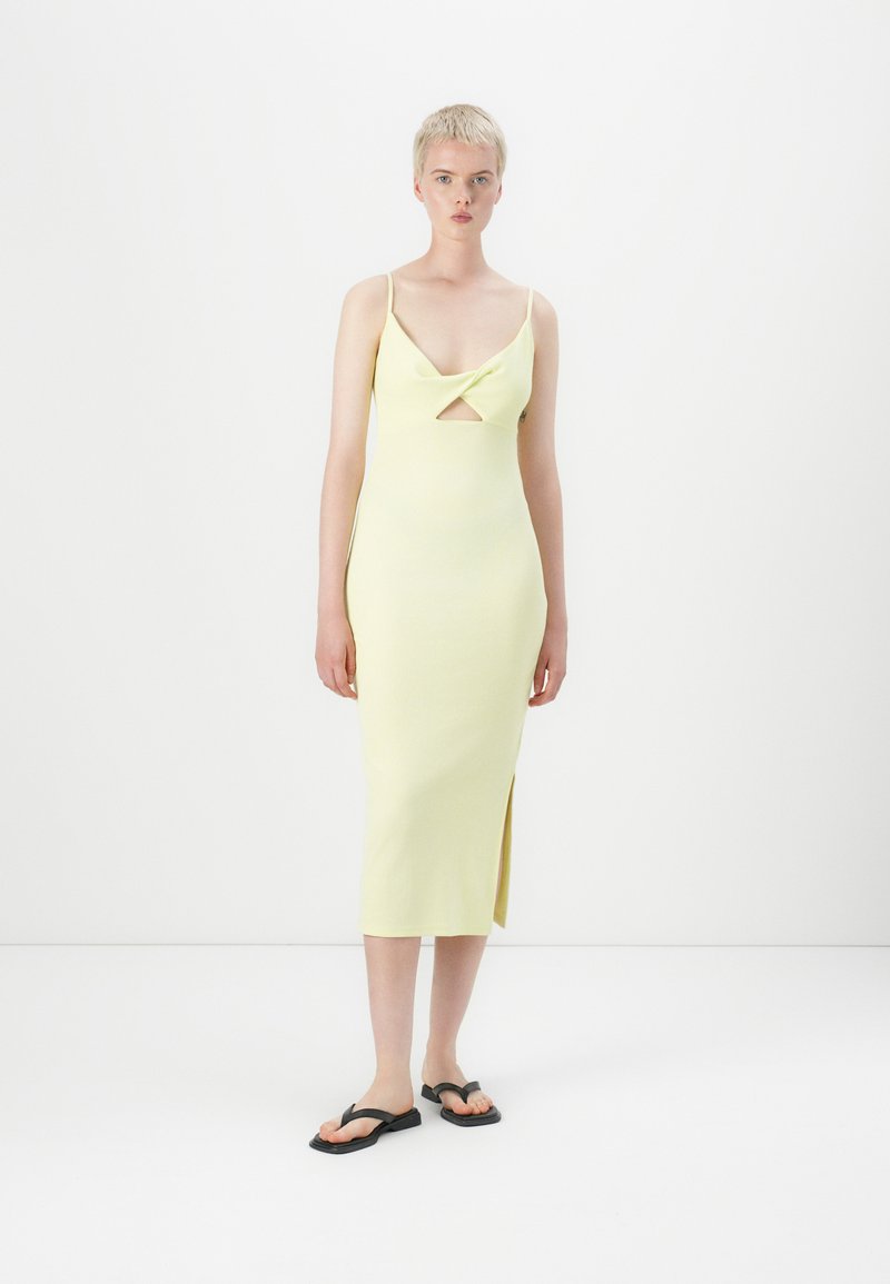 Robe midi en tricot de couleur jaune clair, avec un design en torsion au niveau de la poitrine, des bretelles fines et une fente sur le côté. Le modèle porte des tongs noires.