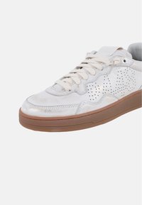 Zapatilla blanca con parte superior de cuero texturizado y perforaciones, cordones en un tono similar y suela de goma marrón. Diseño de punta redondeada.